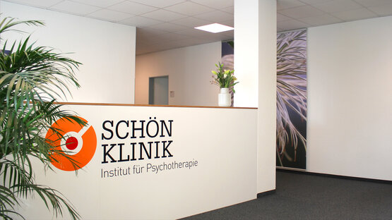 Heller Empfangsbereich mit weißer Theke, Logo „Schön Klinik Institut für Psychotherapie“, Blumen und Pflanzen