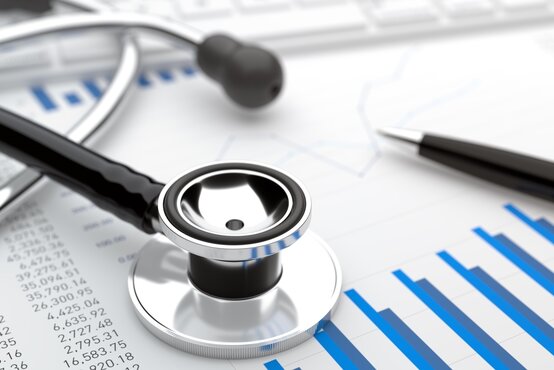 Nahaufnahme eines Stethoskops und eines Kugelschreibers auf einem Blatt mit blauen Balkendiagrammen und medizinischen Statistiken