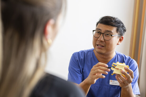 Orthopäde im blauen Kittel mit Namensschild erklärt im Rückeninstitut einer Patientin mit Wirbelsäulenmodell Rückenprobleme