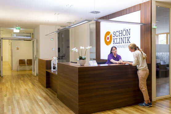 Moderner Empfangsbereich des Schön Klinik Rückeninstituts mit Holzfußboden, Empfangstresen, beleuchtetem Logo und zwei Personen im Gespräch