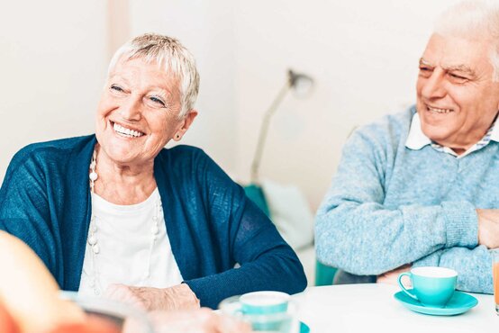 Zwei ältere Menschen sitzen an einem Tisch mit blauen Tassen und Saftglas, beide lachen