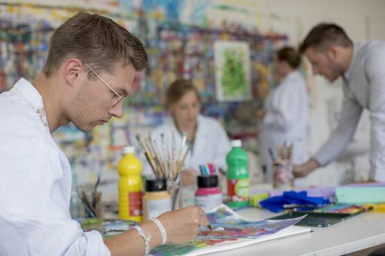 Mehrere Personen in weißen Kitteln malen gemeinsam an einem Tisch mit Farben und Pinseln in einem Kunstraum, farbenfrohes Bild an der Wand