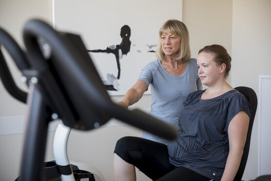 Junge Frau sitzt auf Trainingsgerät, Therapeutin erklärt Bedienung in hellem Therapieraum