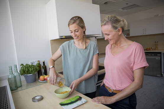 Zwei Frauen bereiten gemeinsam Frühstück in heller Therapieküche zu, Cornflakes werden in grüne Schüssel gefüllt