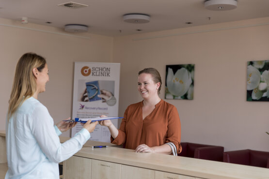 Zwei Frauen am Empfangstresen, eine übergibt der anderen ein Klemmbrett, Roll-up „Schön Klinik“ im Hintergrund