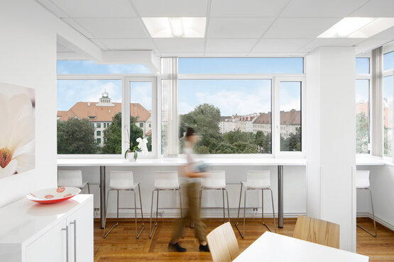 Helle Aufenthaltsraum der Tagesklinik München mit weißen Möbeln, Barhockern an Fensterfront, Panoramablick auf Stadt mit roten Dächern