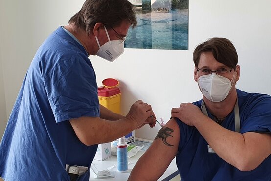 Zwei männliche Klinikmitarbeiter in blauer Arbeitskleidung und FFP2-Masken, Covid-19-Impfung wird geimpft