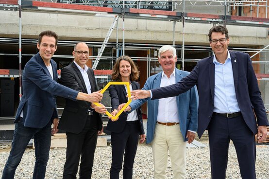 Fünf Personen in Businesskleidung halten gemeinsames gelbes Symbol und lächeln vor Baustelle des Ronald McDonald Hauses Vogtareuth