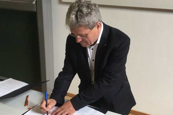 Älterer Mann im Anzug unterschreibt Vertrag an Tisch im Büro, Unterlagen und Kugelschreiber liegen bereit, Szene zur Vertragsunterzeichnung