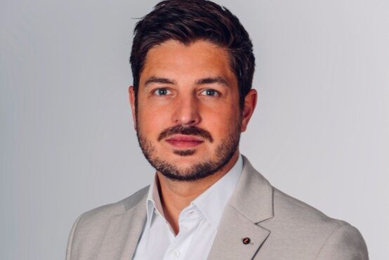 Porträt von einem Mann im hellen Sakko und weißen Hemd mit kurzem dunklem Haar und Bart vor neutralem Hintergrund