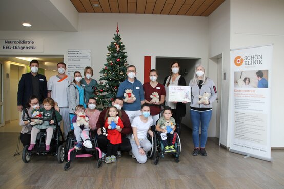 Gruppenfoto mit Kindern im Rollstuhl, Fachpersonal mit Masken, Stofftieren und Weihnachtsbaum