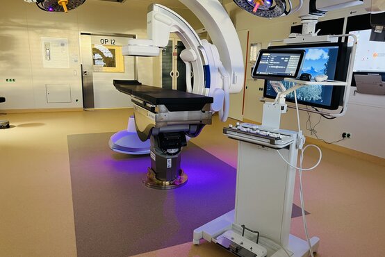 Hybrid-OP-Saal mit verstellbarem OP-Tisch, integriertem C-Bogen, Steuerpult mit Monitoren, modernen LED-OP-Leuchten und Tür „OP 12“