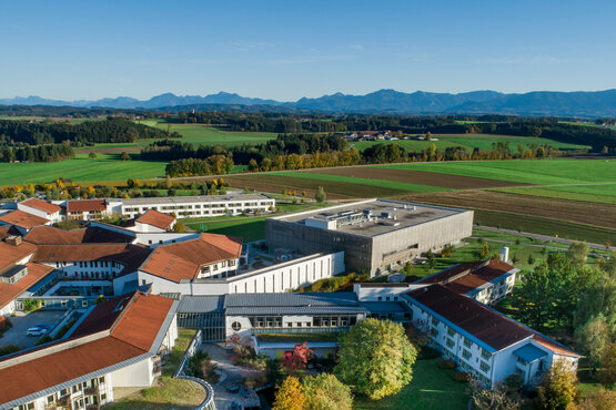 Luftaufnahme der Schön Klinik Vogtareuth mit modernen Gebäuden, roten Ziegeldächern, begrünten Höfen und Alpenpanorama im Hintergrund