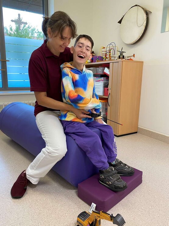 Lächelnder Junge sitzt auf blauer Gymnastikrolle, Therapeutin im roten Shirt unterstützt ihn im hellen Therapieraum 