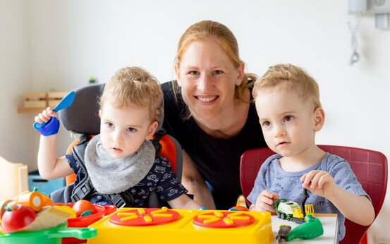 Lächelnde Therapeutin sitzt mit zwei kleinen Jungen an Kliniktisch beim Spielen mit buntem Lernspielzeug und Küchenutensilien