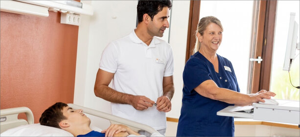 Helles Klinikzimmer mit jungem Patienten im Bett, zwei Fachkräfte – Mann in Weiß und Frau in dunkelblauer Pflegekleidung – am Computer