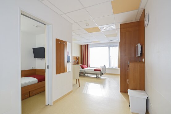 Familienzimmer auf der Kinderstation mit Klinikbett, Einzelbett im Nebenraum, Fensterfront, roter Vorhang und medizinischer Ausstattung