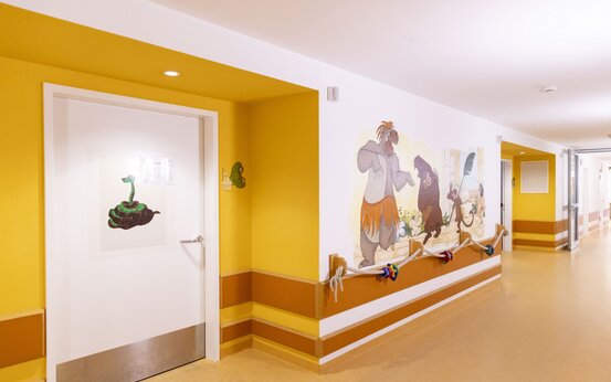 Klinik Flur mit breitem Handlauf, bunten Motorik-Spielzeugen, Dschungelbuch-Wandbild und farbiger Tür für kinderfreundliche Therapieumgebung