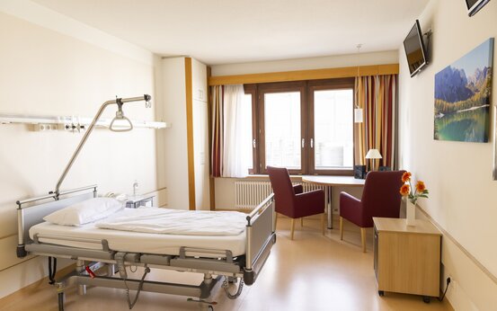 Modernes Einzel-Patientenzimmer mit höhenverstellbarem Klinikbett, Fenster, Sitzbereich und Holzboden