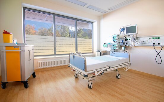 Modernes, helles Klinik-Patientenzimmer mit großem Fenster, Krankenhausbett, medizinischer Gerätewand und gelbem Notfallwagen
