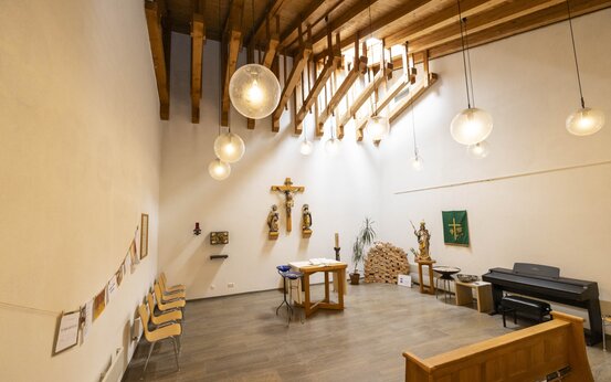 Kapelle mit hoher Holzdecke, Kreuzigungsdarstellung, Holzaltar, gepolsterten Stühlen, E-Piano und freiem Zugang in Klinik