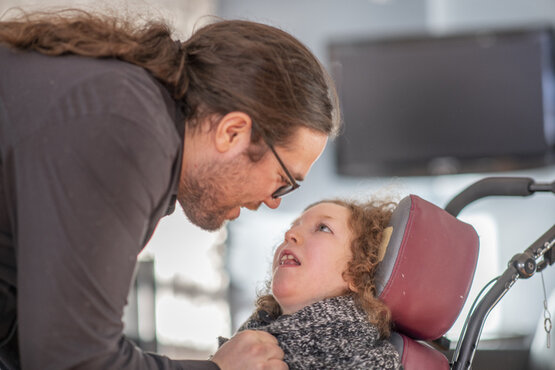 Erwachsene mit Brille beugt sich zu Kind mit lockigem Haar im roten Rollstuhl und gestricktem Schal