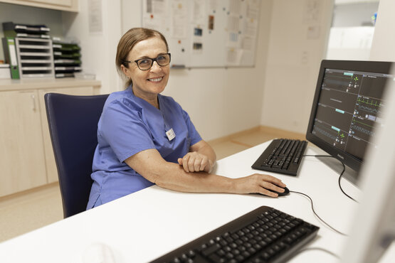 Pflegekraft in blauer Klinikkleidung arbeitet an Computer mit Überwachungsmonitoren an modernem Schreibtisch in Klinikbüro