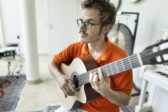 Mann mit Brille und orangefarbenem Poloshirt spielt akustische Gitarre in hellem Musikraum mit Schlagzeug, Gong und Keyboard