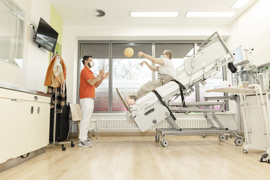 Patient sitzt mit Maske auf aufgerichtetem Klinikbett und spielt Therapieball zu Therapeut im orangefarbenen Shirt