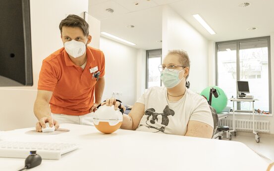  Therapeut in orangem Poloshirt betreut Patientin mit Maske bei Handtraining mit Therapiegerät am Tisch