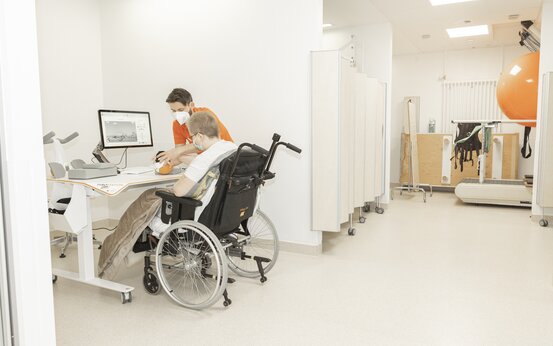 Patient im Rollstuhl und Therapeutin mit Maske trainieren Handmotorik am Therapiegerät mit Computer im modernen, hellen Therapieraum