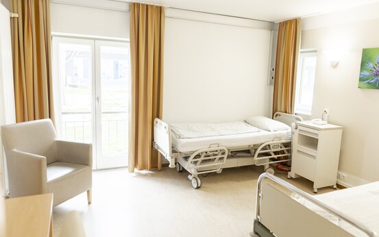 Patientenzimmer in der Schön Klinik Vogtareuth mit höhenverstellbarem Pflegebett, Nachttisch, Besuchersessel und großem Fenster