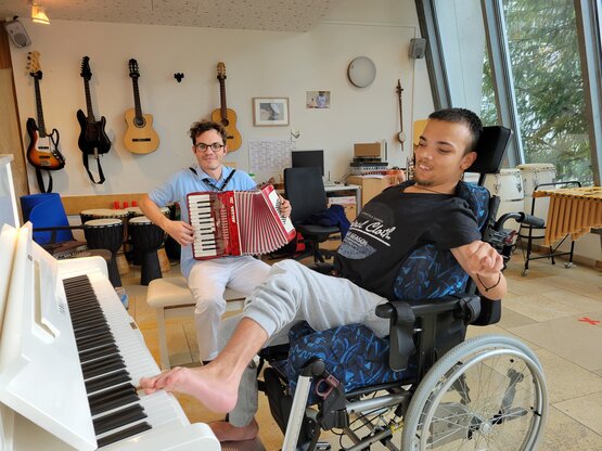 Junger Mann im Rollstuhl spielt barfuß mit dem Fuß Klavier, Musiktherapeut begleitet aufmerksam mit Akkordeon