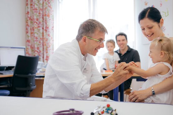Arzt im weißen Kittel begrüßt lächelnd ein kleines Kind auf dem Arm einer Frau im hellen Untersuchungszimmer, Familie im Hintergrund