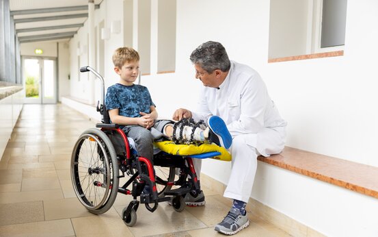 Arzt im weißen Kittel begutachtet Bein eines Kindes mit Fixateur und gelagertem Bein im Rollstuhl