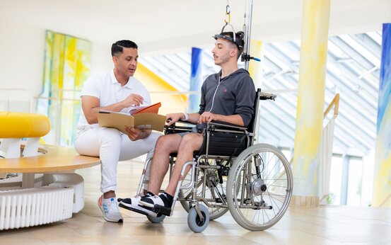 Mitarbeiter in Klinik bespricht Akte mit jungem Mann im Rollstuhl mit Kopfhalterung