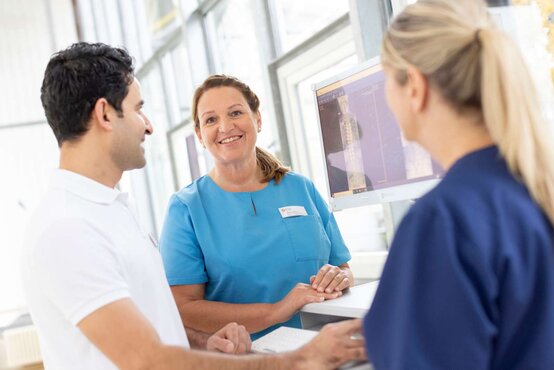 Drei medizinische Fachkräfte am Schreibtisch in heller Praxis, Frau in Blau lächelt in die Kamera, Bildschirm mit Wirbelsäulenaufnahme