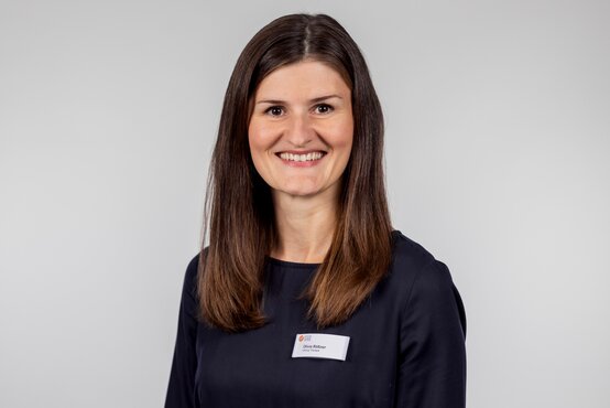Julia Rößner, lächelt freundlich mit Namensschild im dunkelblauen Oberteil vor neutralgrauem Hintergrund im Klinik-Porträt
