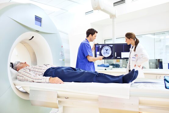 Patient liegt zur CT-Untersuchung im Scanner, zwei Fachkräfte betrachten CT-Bilder vom Gehirn auf Monitoren
