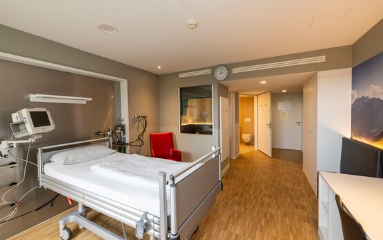 Komfort-Patientenzimmer der Schön Klinik Vogtareuth mit Pflegebett, rotem Sessel, Schrank, Schreibtisch und Badezimmer