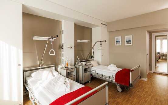 Klinikzimmer mit zwei Pflegebetten, Nachttischen, Aufrichthilfen, Holzfußboden und heller, freundlicher Atmosphäre