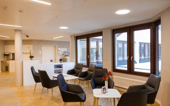 Komfort-Lounge der Schön Klinik Vogtareuth mit bequemen Sesseln, weißen Tischen, Parkettboden, großen Fenstern, Empfangsbereich, Zeitschriften und Kaffee