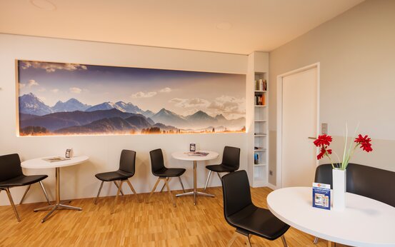 Helle Komfort-Lounge der Schön Klinik Vogtareuth mit großem Bergpanorama Bild, Holzfußboden, runden weißen Tischen und gepolsterten Stühlen