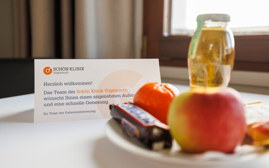 Willkommenskarte der Schön Klinik Vogtareuth, weißer Teller mit Apfel, Mandarine, Keks und Schokoriegel sowie Glas Saft auf weißem Tisch