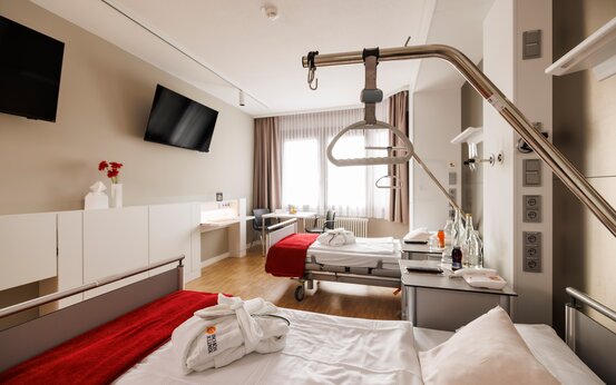 Komfort-Doppelzimmer der Schön Klinik Vogtareuth mit zwei Pflegebetten, Nachttischen, großem Fenster, Flachbildfernsehern, Schreibtisch und Holzboden