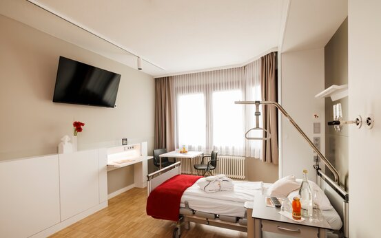 Einzelzimmer der Schön Klinik Vogtareuth mit Pflegebett, Nachttisch, Flachbild-TV, Schreibtisch mit Blume, Sitzecke und Fenstern