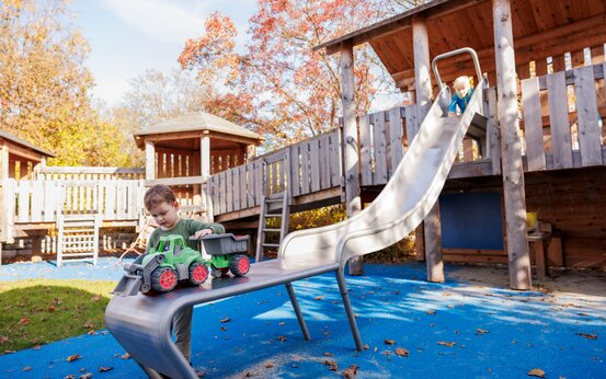 Bunter Klinikspielplatz mit Klettergerüst und Rutsche, Junge spielt mit Trecker am Ende der Rutsche, ein anderer steht am Start der Rutsche