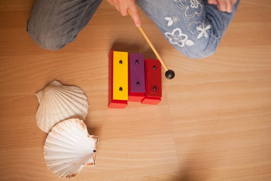 Kind in blauer Jeans mit Blumenmuster sitzt auf hellem Holzboden und spielt mit buntem Xylophon und Muschelschalen in Musiktherapie