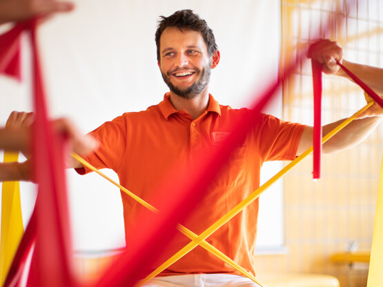 Therapeut im orangefarbenen Poloshirt hält gelbes und rotes Therapieband, weitere gespannte Bänder und Arme im Vordergrund
