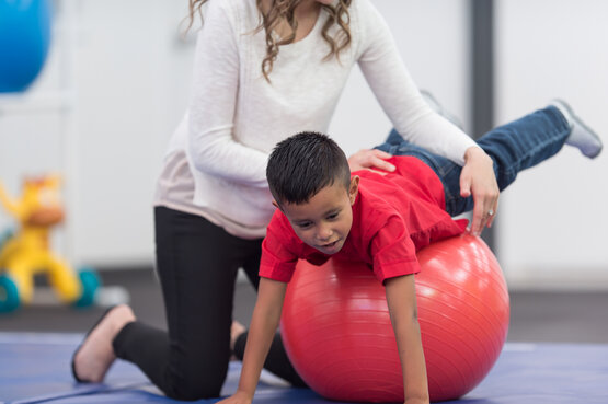 Kind liegt bäuchlings auf rotem Gymnastikball, Therapeutin sichert mit beiden Händen
